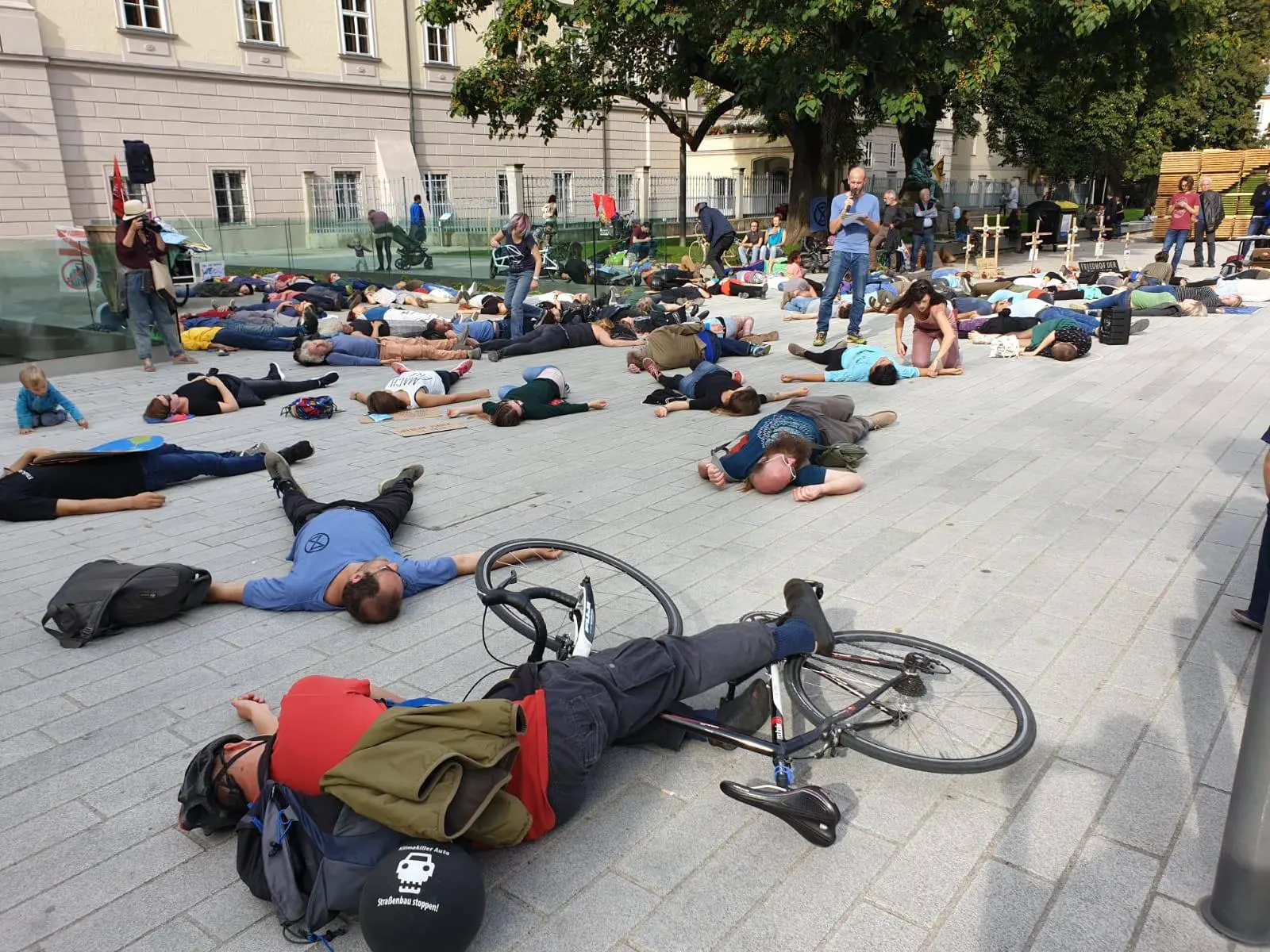 AktivistInnen und SympatisantInnen von XR Oberösterreich liegen vor dem Landhaus am Boden. Ein Aktivist hält eine Rede. Das Die in symbolisiert das Artensterben und die Bedrohung durch die Klimakrise